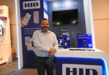 HID impulsa las identidades seguras con un ecosistema que transforma el control de acceso Fotografía de un hombre sonriente, Luis Yáñez, con camisa celeste y pantalón oscuro, de pie frente a un stand de exhibición de la marca HID. El módulo, iluminado con luces LED azules, muestra letreros promocionales sobre alianzas tecnológicas y exhibe dispositivos de seguridad como un lector de reconocimiento facial (HID Amico) y un lector de acceso móvil (HID Mobile Access) sobre un mostrador blanco.