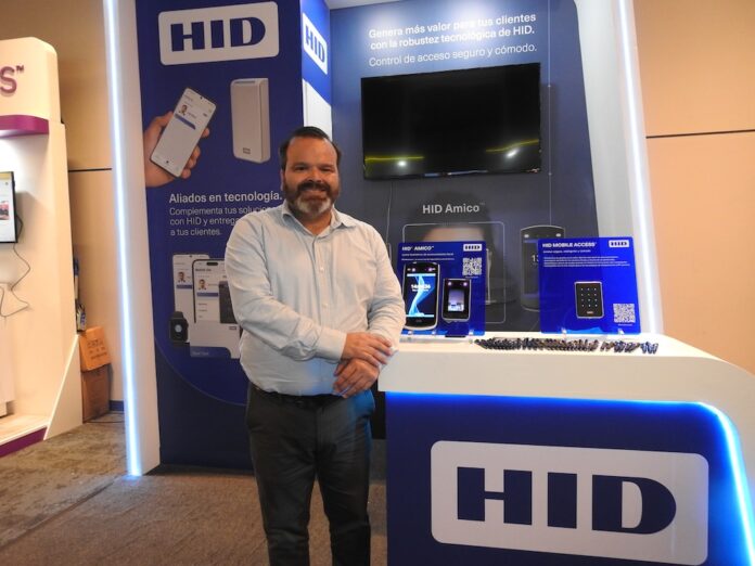 La revolución de las identidades seguras. De la tarjeta física al ecosistema digital móvil Fotografía de un hombre sonriente, Luis Yáñez, con camisa celeste y pantalón oscuro, de pie frente a un stand de exhibición de la marca HID. El módulo, iluminado con luces LED azules, muestra letreros promocionales sobre alianzas tecnológicas y exhibe dispositivos de seguridad como un lector de reconocimiento facial (HID Amico) y un lector de acceso móvil (HID Mobile Access) sobre un mostrador blanco.