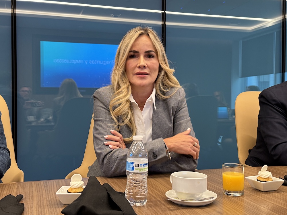 Fotografía de medio cuerpo de Laura Gómez, sentada en una sala de conferencias durante un encuentro con medios. La ejecutiva viste un saco gris sobre una camisa blanca y mantiene los brazos cruzados apoyados sobre una mesa de madera. En primer plano hay una botella de agua de plástico, una taza de café blanca, un vaso con jugo de naranja y dos pequeños bocadillos. Al fondo, detrás de un panel de cristal azul, se percibe una pantalla proyectando el texto "Preguntas y respuestas".