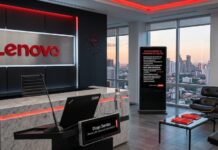 Lenovo recompensa la lealtad de sus clientes con su nuevo programa Puntos Lenovo Fotografía de una moderna oficina corporativa con acabados de lujo. En primer plano hay un escritorio de mármol con un micrófono, una laptop y una placa digital que dice "Diego Darriba". En la pared oscura detrás del escritorio resalta el logotipo tridimensional de Lenovo iluminado en rojo. A la derecha, un letrero digital vertical promociona "PUNTOS LENOVO: TU PROGRAMA DE LEALTAD" junto a una sala de espera con sillas de cuero negro y grandes ventanales que muestran la ciudad al atardecer.