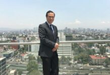 Alejandro Apan, director general del Neocenter: “Los espacios de colaboración apenas han cubierto el 30% del mercado” Retrato de medio cuerpo de Alejandro Apan, Director General de Neocenter, de pie en una terraza corporativa. Viste un traje oscuro bien cortado, una camisa de cuadros finos y una corbata de seda azul marino. Tiene las manos entrelazadas al frente y sonríe ligeramente a la cámara. El fondo muestra una vista panorámica de la Ciudad de México bajo un cielo despejado y brillante, con edificios de oficinas y residencias que se extienden hasta el horizonte.