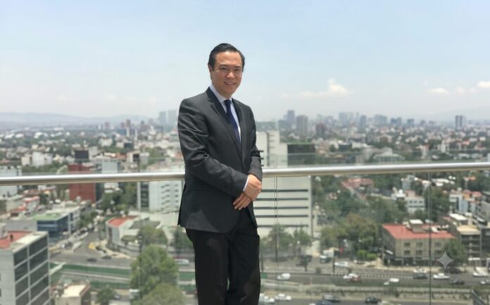 Retrato de medio cuerpo de Alejandro Apan, Director General de Neocenter, de pie en una terraza corporativa. Viste un traje oscuro bien cortado, una camisa de cuadros finos y una corbata de seda azul marino. Tiene las manos entrelazadas al frente y sonríe ligeramente a la cámara. El fondo muestra una vista panorámica de la Ciudad de México bajo un cielo despejado y brillante, con edificios de oficinas y residencias que se extienden hasta el horizonte.