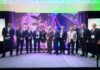 NETSCOUT analiza el impacto de la inteligencia artificial en la ciberseguridad y las operaciones de TI Fotografía grupal de once profesionales posando de pie sobre un escenario alfombrado; el grupo está conformado por diez hombres de traje y una mujer al centro con vestido negro y detalles blancos. El fondo muestra una gran mampara iluminada con luces ambientales en tonos verdes y morados que dice "NETSCOUT Partner Summit".