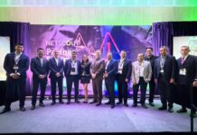 NETSCOUT analiza el impacto de la inteligencia artificial en la ciberseguridad y las operaciones de TI Fotografía grupal de once profesionales posando de pie sobre un escenario alfombrado; el grupo está conformado por diez hombres de traje y una mujer al centro con vestido negro y detalles blancos. El fondo muestra una gran mampara iluminada con luces ambientales en tonos verdes y morados que dice "NETSCOUT Partner Summit".