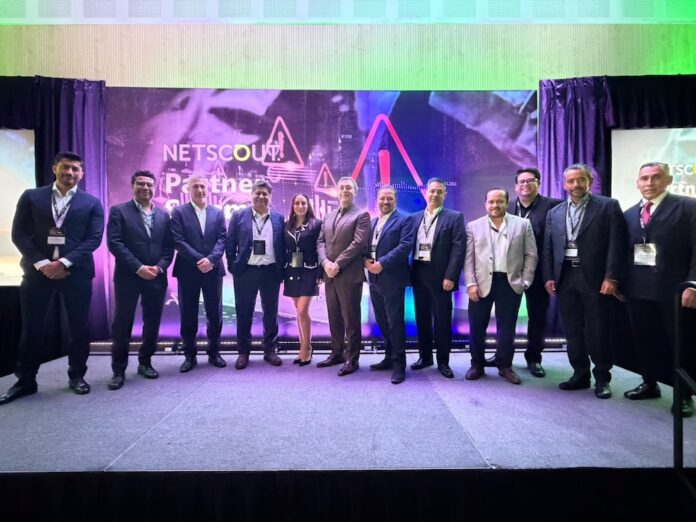 Fotografía grupal de once profesionales posando de pie sobre un escenario alfombrado; el grupo está conformado por diez hombres de traje y una mujer al centro con vestido negro y detalles blancos. El fondo muestra una gran mampara iluminada con luces ambientales en tonos verdes y morados que dice 