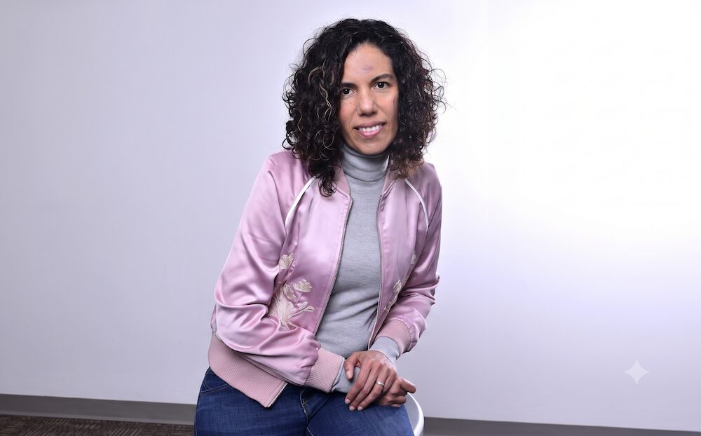 Retrato en plano medio de Lorena Escandón, directora de Comunicación y Marketing en Xerox, posando sentada con el cuerpo ligeramente inclinado de tres cuartos frente a un fondo blanco. La ejecutiva, de tez clara y cabello oscuro rizado con luces doradas, sonríe a la cámara vistiendo un suéter gris de cuello alto y una chamarra deportiva satinada de color rosa claro con detalles bordados de flores blancas y bordes elásticos en color rosa pálido, manteniendo las manos entrelazadas sobre su pierna.