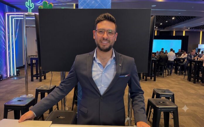 Retrato de medio cuerpo de Luis Fernando Celis, hombre con gafas, barba corta y un saco azul oscuro sobre camisa celeste, posando sonriente detrás de un mostrador en un salón de eventos corporativo con asistentes al fondo.