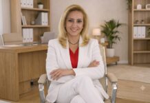 TechZone POS impulsa la capacitación del canal con certificaciones gratuitas en soluciones para punto de venta Fotografía en plano medio que captura a Maru Navas, Gerente Senior de Canales en TechZone, sentada de frente en una silla de oficina ergonómica de color beige con descansabrazos cromados. La ejecutiva, de tez clara y cabello rubio lacio de largo medio, sonríe a la cámara vistiendo un conjunto formal de saco y pantalón blancos sobre una blusa interior color rojo vivo, complementado con una gargantilla delgada negra. Al fondo, de manera difuminada, se observa un entorno de oficina moderno y luminoso con muebles de madera clara, un escritorio con laptop, estantes con carpetas blancas archivadoras y plantas decorativas.