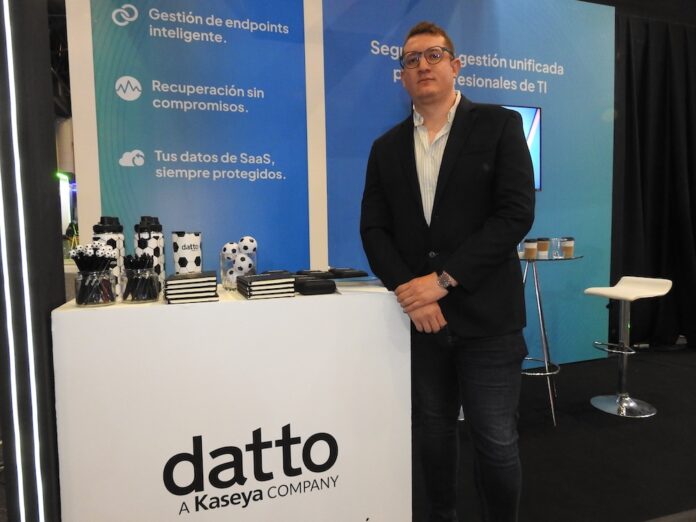 Mateo Bolívar detalla el impacto de la automatización en la ciberseguridad corporativa Retrato de Mateo Bolívar, gerente comercial de nuevos negocios para Latinoamérica en Kaseya, quien posa de frente con las manos entrelazadas a la altura de la cintura. El ejecutivo usa anteojos y viste un saco oscuro sobre una camisa clara y jeans. Se encuentra de pie junto a un mostrador blanco de la marca Datto, respaldado por un panel informativo azul que destaca beneficios como