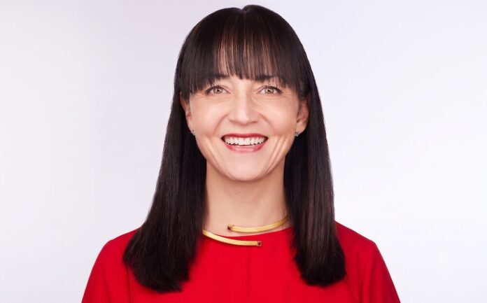 Retrato de estudio en plano medio y de frente de Mika Yamamoto, directora de Marketing (CMO) y de IA para el cliente de Veeam Software, posando sonriente sobre un fondo blanco limpio. La ejecutiva tiene tez clara, cabello oscuro y lacio con fleco recto que enmarca su rostro, y viste una blusa formal de color rojo brillante. En su cuello resalta una gargantilla rígida dorada con un diseño asimétrico minimalista y discretas incrustaciones cuadradas de pedrería roja en los extremos.
