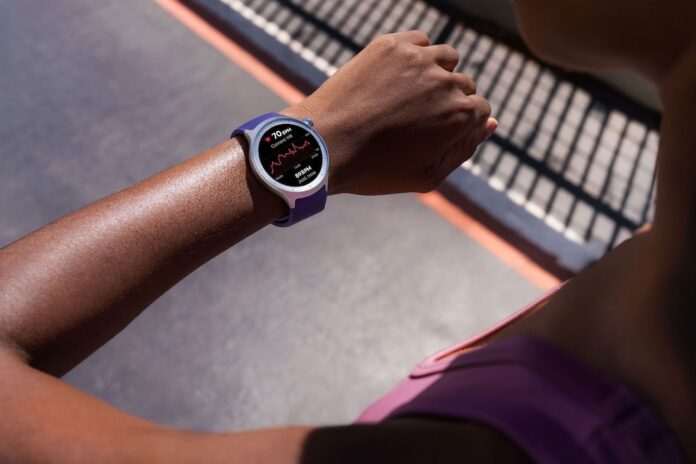 Fotografía en primer plano del brazo de una persona vistiendo ropa deportiva al aire libre bajo la luz del sol, llevando en la muñeca un moto watch de caja plateada y correa color morado. La pantalla circular del reloj inteligente está encendida con fondo negro y muestra una gráfica roja de frecuencia cardíaca en tiempo real, indicando la lectura 