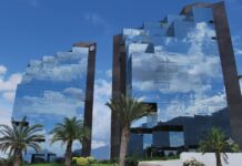 Grupo CVA fortalece su presencia en Monterrey con una nueva sucursal para acelerar el crecimiento del canal TI Fotografía exterior de dos imponentes edificios corporativos con diseño arquitectónico escalonado y fachadas completamente recubiertas de cristales, los cuales reflejan un cielo azul con nubes blancas y la silueta de las montañas. En primer plano se aprecian palmeras y jardines verdes bien cuidados.