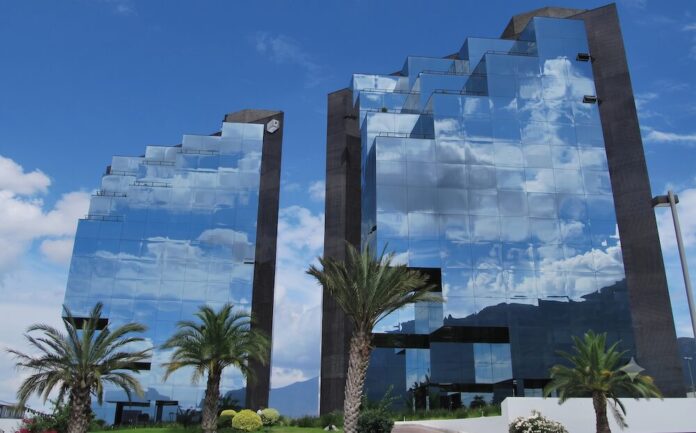 Nueva sucursal de Grupo CVA en Monterrey. Evolución y cercanía corporativa Fotografía exterior de dos imponentes edificios corporativos con diseño arquitectónico escalonado y fachadas completamente recubiertas de cristales, los cuales reflejan un cielo azul con nubes blancas y la silueta de las montañas. En primer plano se aprecian palmeras y jardines verdes bien cuidados.