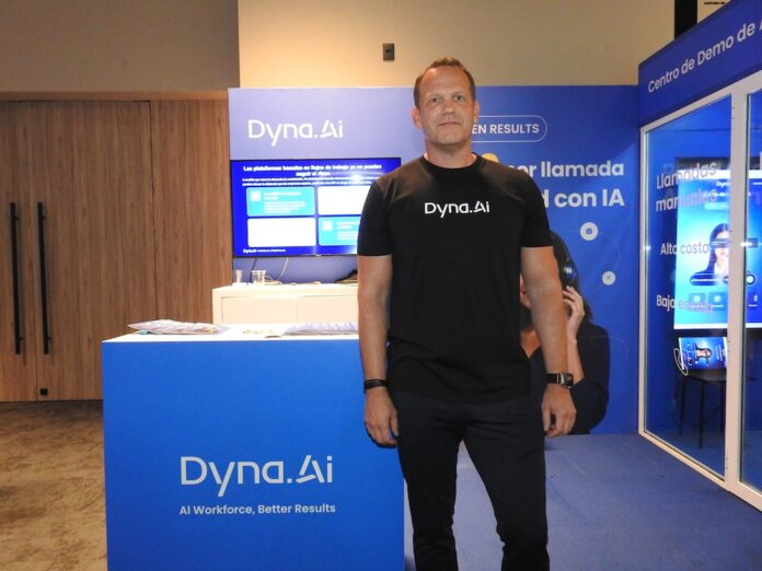 Peter Dallen analiza la reducción de latencia en sistemas de inteligencia artificial para centros de contacto Fotografía en plano medio que muestra a Peter Dallen, presidente para América Latina de Dyna.Ai, posando de frente con una postura firme y formal en el espacio de exhibición de su compañía. El directivo, de tez clara y complexión robusta, viste una playera negra lisa con el logotipo blanco de