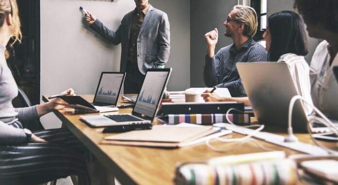 Planificación estratégica y análisis. El camino hacia un cierre de año empresarial exitoso Fotografía de una reunión de negocios donde un hombre de traje gris está de pie señalando un pizarrón blanco. A su alrededor, un equipo diverso de profesionales sonríe y presta atención sentados en una amplia mesa de madera. En primer plano, destacan dos computadoras portátiles con las pantallas abiertas mostrando gráficas financieras de barras y líneas, acompañadas de libretas, bolígrafos y vasos de café.