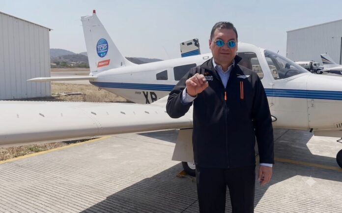 Fotografía en plano medio que muestra a un directivo de telecomunicaciones posando de frente en la pista de hormigón de un aeródromo durante el día. El ejecutivo, que porta gafas de sol con cristales de espejo azul, viste una chamarra ejecutiva negra con cierre que lleva bordados los logotipos de 