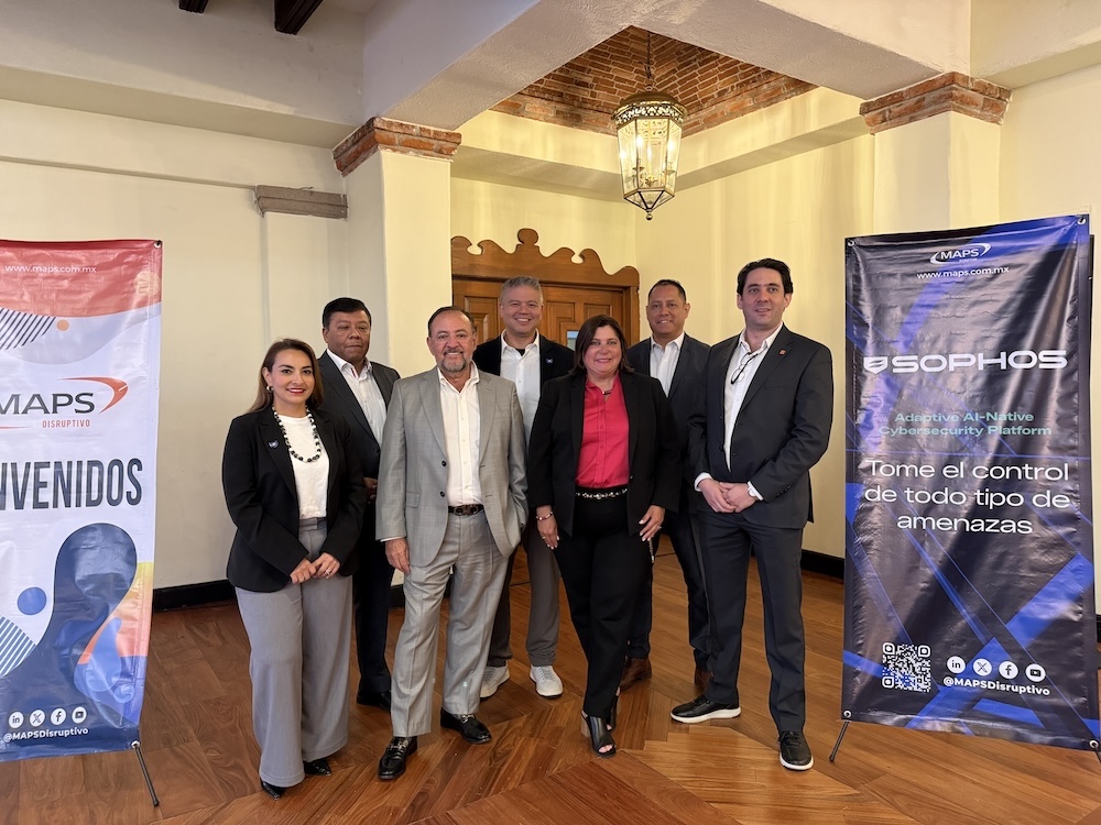 Fotografía en plano general que captura a siete colaboradores y directivos de MAPS y Sophos posando juntos de frente en el salón del evento. El grupo, integrado por cinco hombres y dos mujeres vestidos con trajes formales, sacos ejecutivos y atuendos de negocios en tonos oscuros y grises, se distribuye de manera ordenada en el centro del espacio con pisos de madera clara. El encuadre está flanqueado simétricamente a la izquierda por el tótem promocional blanco y azul de "MAPS DISRUPTIVO" y a la derecha por el banner publicitario negro de "SOPHOS" bajo el lema "Tome el control de todo tipo de amenazas".