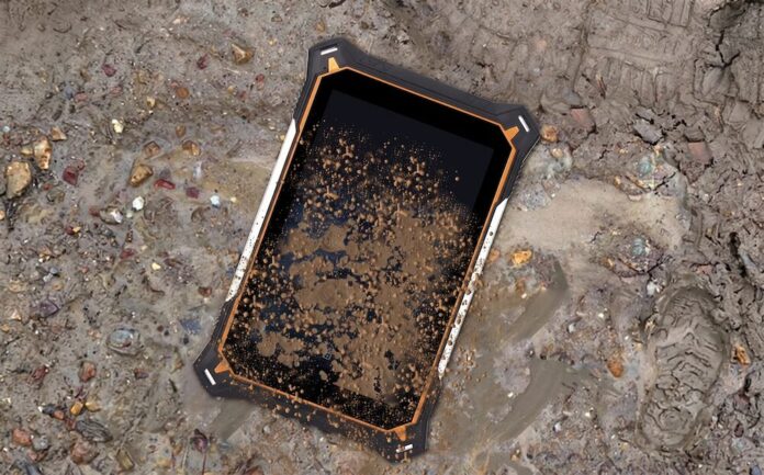 Fotografía de producto en condiciones reales de uso que muestra la pantalla frontal de la tableta Jaeger 8A de ML Rugged recostada de forma diagonal sobre un terreno lodoso, húmedo y con piedras sueltas. La superficie de la pantalla de cristal templado y los bordes con detalles naranjas y blancos del chasis reforzado se encuentran severamente cubiertos por salpicaduras densas de lodo marrón oscuro, mientras que a la derecha, en la tierra mojada, se distingue la huella de una bota de trabajo industrial.