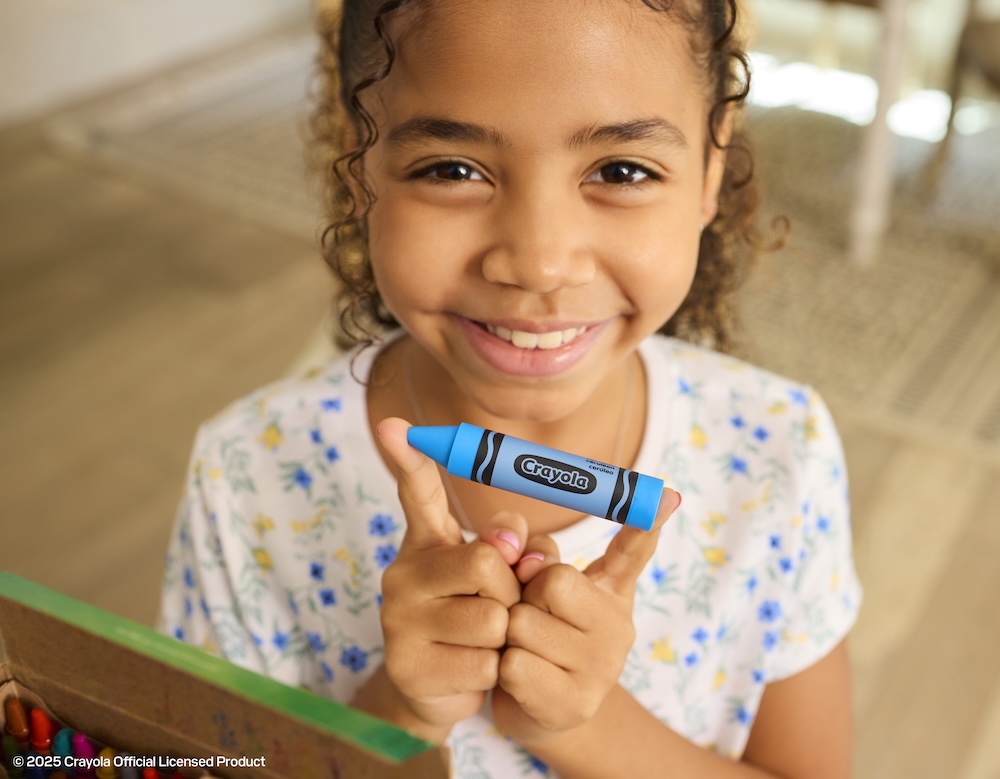 Fotografía en primer plano de una niña sonriendo a la cámara mientras sostiene a la altura de su barbilla una unidad flash USB-C con el diseño exacto de un crayón Crayola en color azul (Cerulean Blue). En la esquina inferior izquierda se asoma ligeramente una caja de crayones tradicional.