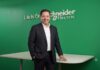 Schneider Electric: El canal TI encuentra un nuevo diferenciador, garantizar la continuidad operativa Fotografía de medio cuerpo de un hombre de negocios sonriente, vestido con un traje oscuro y camisa blanca sin corbata. El sujeto se encuentra de pie, con la mano derecha en el bolsillo y la izquierda apoyada sobre una mesa blanca. Detrás de él, resalta una pared de color verde brillante con el logotipo de la empresa y el lema "Life Is On Schneider Electric" en letras blancas.