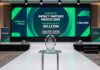 Sellcom obtiene el reconocimiento Impact Partner Mexico 2025 de Veeam Fotografía de un elegante escenario de premiación. En primer plano, un trofeo geométrico de cristal descansa sobre una mesa transparente. De fondo, una gran pantalla iluminada en tonos verdes y azules anuncia en letras blancas: "GANADOR: IMPACT PARTNER MEXICO 2025", "RECONOCIMIENTO A: SELLCOM" y "VEEAM IMPACT PARTNER AWARDS 2025 | AMERICAS". A los costados del escenario alfombrado se observan pendones corporativos con los logotipos de Veeam y Sellcom.
