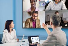 ViewSonic fortalece la ciberseguridad empresarial con su nueva serie CDEG3 de pantallas 4K Fotografía de dos profesionales en una sala de reuniones con paredes azules. Un hombre, visto de espaldas, gesticula con la mano derecha hacia una gran pantalla montada en la pared que muestra una videoconferencia con cuatro participantes. En la mesa blanca frente a él hay una computadora portátil conectada por cable, la cual muestra gráficos y datos en su pantalla. A su izquierda, una mujer con blusa blanca sonríe mientras presta atención a la reunión virtual.