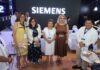 Siemens fortalece la digitalización de las redes eléctricas para acelerar la transición energética Fotografía grupal de cinco personas posando sonrientes frente a un gran letrero luminoso con la marca "SIEMENS". El grupo está conformado por cuatro mujeres y un hombre; todos visten de manera formal-casual, portan gafetes del evento y se encuentran de pie en el área central de la exhibición, rodeados de elementos tecnológicos y mesas de atención.