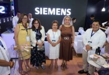 Siemens fortalece la digitalización de las redes eléctricas para acelerar la transición energética Fotografía grupal de cinco personas posando sonrientes frente a un gran letrero luminoso con la marca "SIEMENS". El grupo está conformado por cuatro mujeres y un hombre; todos visten de manera formal-casual, portan gafetes del evento y se encuentran de pie en el área central de la exhibición, rodeados de elementos tecnológicos y mesas de atención.