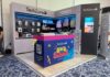 TechZone impulsa el Regreso a Clases 2026 con accesorios inalámbricos llenos de color y productividad Vista de tres cuartos de un stand de exhibición comercial de la marca mexicana TechZone en una convención con piso alfombrado azul. Al frente del stand destaca un mostrador azul y rosa decorado con ilustraciones escolares amarillas y el logotipo "Back to School, ¡Nos movemos contigo!", junto con un código QR para escanear su catálogo completo. En las paredes de fondo gris se exhiben empaques de accesorios y periféricos de cómputo bajo la leyenda "Accesorios y periféricos de alta precisión". De lado derecho, se muestran colgadas tres mochilas escolares en colores azul, rosa y verde pastel, junto a una pantalla vertical que proyecta imágenes de productos de la marca.