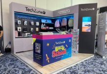 TechZone impulsa el Regreso a Clases 2026 con accesorios inalámbricos llenos de color y productividad Vista de tres cuartos de un stand de exhibición comercial de la marca mexicana TechZone en una convención con piso alfombrado azul. Al frente del stand destaca un mostrador azul y rosa decorado con ilustraciones escolares amarillas y el logotipo "Back to School, ¡Nos movemos contigo!", junto con un código QR para escanear su catálogo completo. En las paredes de fondo gris se exhiben empaques de accesorios y periféricos de cómputo bajo la leyenda "Accesorios y periféricos de alta precisión". De lado derecho, se muestran colgadas tres mochilas escolares en colores azul, rosa y verde pastel, junto a una pantalla vertical que proyecta imágenes de productos de la marca.