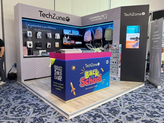 Vista de tres cuartos de un stand de exhibición comercial de la marca mexicana TechZone en una convención con piso alfombrado azul. Al frente del stand destaca un mostrador azul y rosa decorado con ilustraciones escolares amarillas y el logotipo 