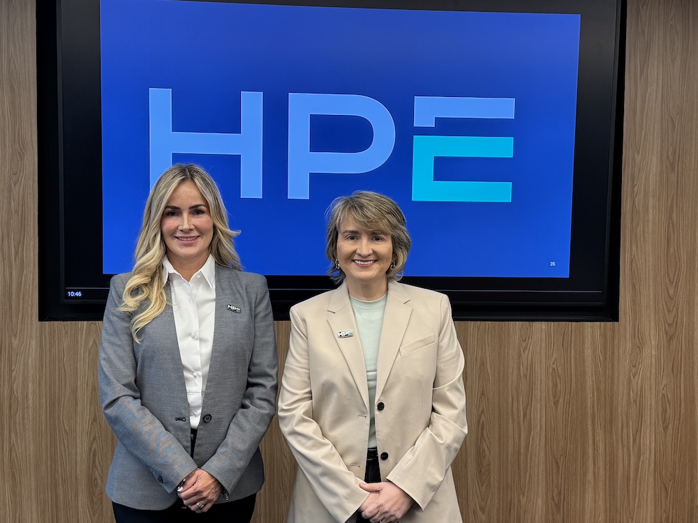 Laura Gómez liderará la estrategia de canales de HPE en México: Fotografía de plano medio de dos ejecutivas de pie, posando sonrientes mirando a la cámara. Detrás de ellas hay una pantalla grande montada en una pared de paneles de madera que proyecta el logotipo de HPE en letras celestes sobre un fondo azul vibrante. A la izquierda se encuentra Laura Gómez, con cabello largo y rubio, vistiendo un saco gris sobre una camisa blanca. A la derecha está Pamela González, con cabello castaño corto, vistiendo un saco beige sobre una blusa verde claro. Ambas ejecutivas portan un pequeño pin con el logotipo de HPE en la solapa izquierda de sus sacos.