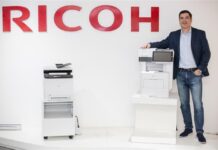 Ricoh apuntala su oferta para partners con “AV Managed Services” Fotografía de Jorge Cortés, manager director para Ricoh, posando de pie con ropa casual de negocios junto a dos impresoras multifuncionales blancas exhibidas bajo el gran logotipo rojo de la marca en la pared.