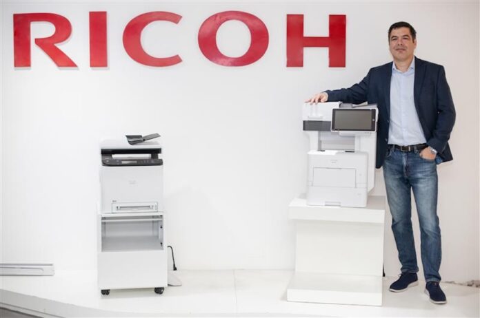 Fotografía de Jorge Cortés, manager director para Ricoh, posando de pie con ropa casual de negocios junto a dos impresoras multifuncionales blancas exhibidas bajo el gran logotipo rojo de la marca en la pared.