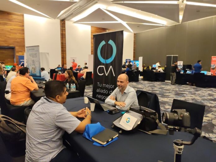 Acompañamiento y desarrollo de proyectos para el canal de TI Fotografía de Zeferino Pérez sentado en una mesa conversando con un asistente dentro de un área de exhibición tecnológica. A su lado se observa un banner promocional vertical color negro con el logotipo de CVA.
