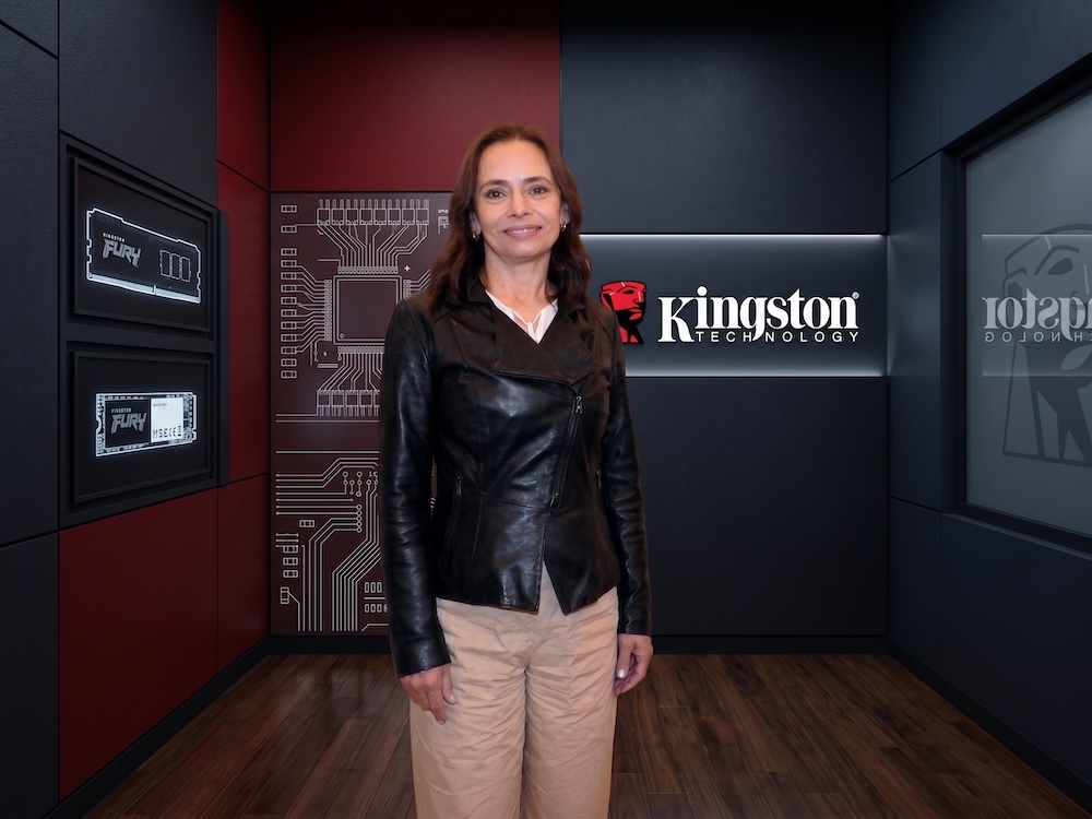 Almacenamiento Kingston: Fotografía de Cristina Carbajal posando de frente y sonriendo a la cámara. Viste una chamarra de cuero negra y pantalones color beige. Al fondo se observa una habitación de exhibición con paredes decoradas con gráficos de circuitos integrados, memorias enmarcadas y el logotipo iluminado de Kingston Technology.
