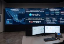 Fortinet y Alestra aceleran la ciberseguridad proactiva con automatización Fotografía del interior de un centro de operaciones de seguridad. En la pared frontal destaca un gran panel de pantallas con los logotipos de Alestra y Fortinet bajo el título "Hub Operacional de Ciberseguridad Avanzada". Los monitores laterales muestran un mapa de conexiones de red en México, gráficas comparativas de activación de VPN y un esquema de ecosistema de seguridad unificado. En primer plano, sobre un escritorio, se observan dos monitores de computadora, un teclado y un teléfono corporativo.