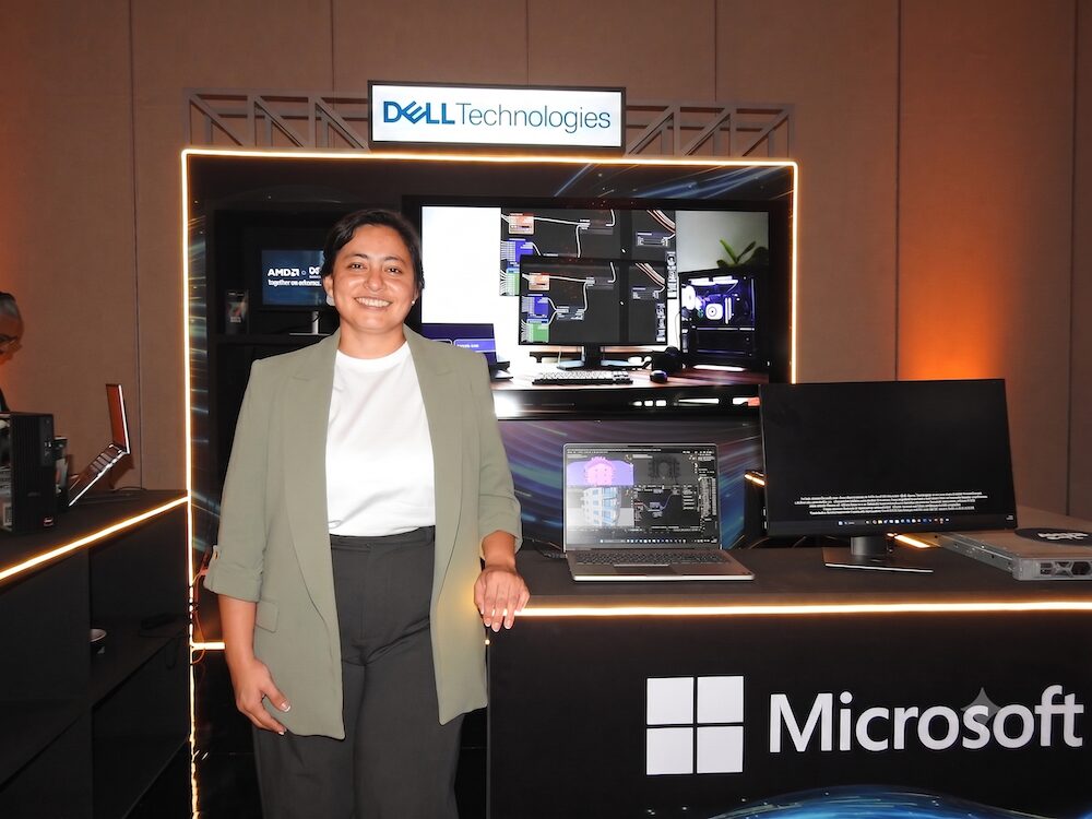 Mujer sonriente vistiendo un saco verde claro, de pie en el stand de Dell Technologies. A su lado, sobre un mostrador oscuro con el logo de Microsoft y AMD, se observan laptops y monitores exhibiendo software de modelado 3D.