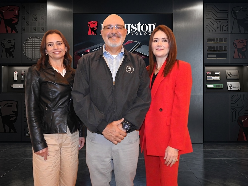 Alianzas en el canal para navegar un mercado de alta exigencia Fotografía grupal de tres ejecutivos posando juntos en un entorno corporativo de exhibición tecnológica. De izquierda a derecha: Cristina Carbajal con chamarra negra, Oscar Martínez al centro con chamarra gris oscura, e Ivette Barragán vistiendo un traje rojo brillante, todos frente a vitrinas y pantallas de productos Kingston.