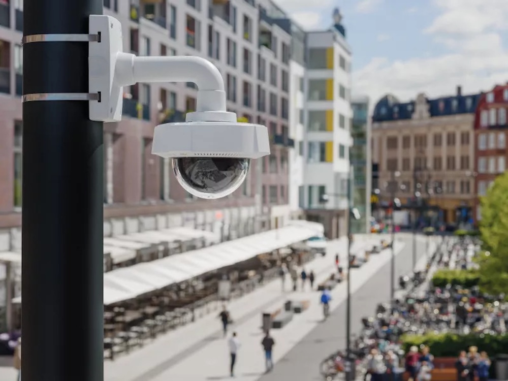 Fotografía de una cámara de videovigilancia blanca tipo domo montada sobre un poste metálico negro. En el fondo, desenfocado, se observa una calle peatonal urbana y soleada con transeúntes, bicicletas estacionadas, terrazas comerciales y edificios de arquitectura clásica y moderna.