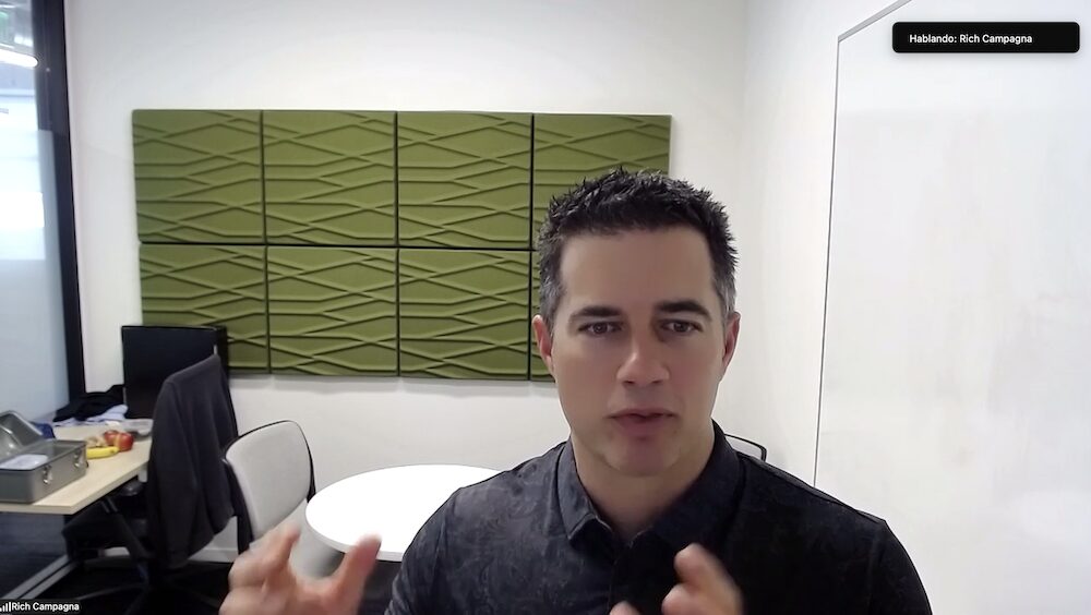 Captura de pantalla de Rich Campagna durante una videoconferencia, hablando y gesticulando levemente con las manos. Se encuentra en una oficina bien iluminada, decorada con paneles acústicos verdes en la pared, una pequeña mesa blanca y un escritorio al fondo.