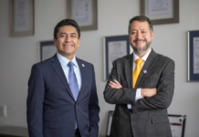 Intelisis transforma el ERP empresarial con IA y automatización Fotografía conjunta de Ángel Baños Carrillo y Julio Olmos posando uno al lado del otro. Ambos ejecutivos visten traje y corbata, y sonríen a la cámara de pie frente a una pared de tonos claros adornada con diplomas enmarcados.