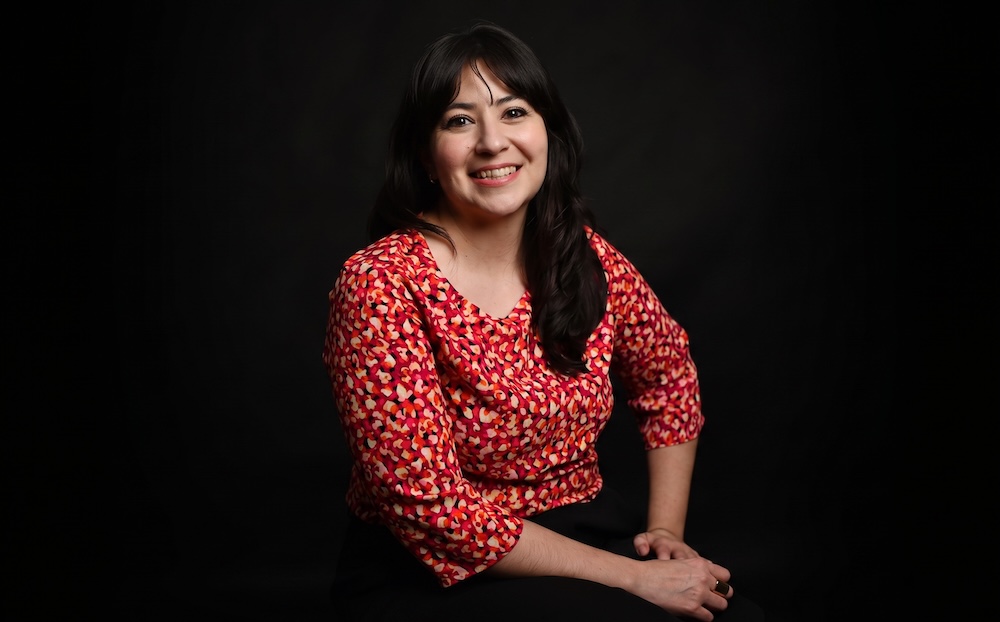Retrato de Tania Álvarez, analista de Select, sonriendo hacia la cámara mientras viste una blusa con estampado en tonos rojos y naranjas sobre un fondo completamente negro.