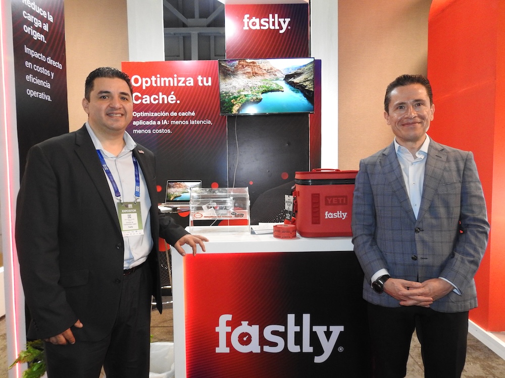 Fotografía de dos hombres sonriendo de pie frente a un stand corporativo de colores rojo, blanco y negro. El hombre a la izquierda usa un gafete y traje oscuro; el de la derecha viste un saco de cuadros grises. El stand muestra logotipos de Fastly y textos sobre optimización de caché, y cuenta con un monitor, una urna de acrílico y una hielera roja de regalo.