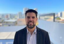 Motorola Solutions fortalece su ecosistema de canales para impulsar nuevos negocios Fotografía de retrato de Arnaldo Lovera. Es un hombre con barba corta y cabello oscuro, que sonríe directamente a la cámara. Viste un saco oscuro sobre una camisa clara abotonada. El fondo muestra un paisaje urbano desenfocado con varios edificios bajo un cielo azul iluminado por el sol.