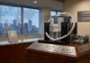 Object First crece 148 % y fortalece su liderazgo en respaldo inmutable contra ransomware Fotografía de una exhibición corporativa ubicada en una oficina moderna. En el centro, sobre una base de madera, destaca un servidor diseñado con la forma de un castillo fortificado que lleva el logotipo de Object First. En la parte inferior, varias placas de metal grabadas muestran estadísticas financieras. De fondo, a través de un ventanal con un mapa del mundo punteado impreso en el cristal, se observa el horizonte de una ciudad al atardecer.