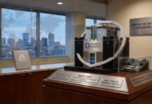 Object First crece 148 % y fortalece su liderazgo en respaldo inmutable contra ransomware Fotografía de una exhibición corporativa ubicada en una oficina moderna. En el centro, sobre una base de madera, destaca un servidor diseñado con la forma de un castillo fortificado que lleva el logotipo de Object First. En la parte inferior, varias placas de metal grabadas muestran estadísticas financieras. De fondo, a través de un ventanal con un mapa del mundo punteado impreso en el cristal, se observa el horizonte de una ciudad al atardecer.