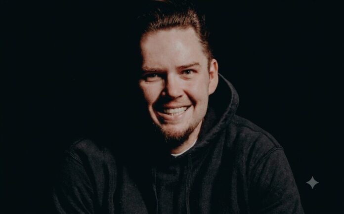 El desafío de la IA generativa en la verificación de identidad Retrato fotográfico de Kris Galloway sonriendo directamente a la cámara. Su rostro está iluminado frontalmente, creando un fuerte contraste con el fondo completamente oscuro, y viste una sudadera negra con capucha.