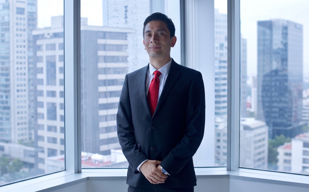 Fotografía de Alejandro Vargas, analista de Select, vistiendo un traje oscuro y corbata roja, posando de pie con las manos cruzadas frente a una ventana con vista a edificios corporativos.