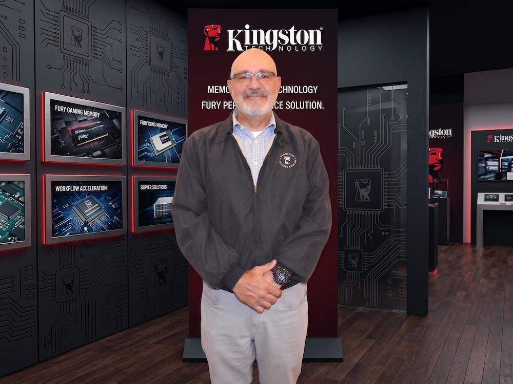Fotografía de retrato de Oscar Martínez, con anteojos y barba, vistiendo una chamarra gris oscura sobre una camisa clara y pantalones grises. Se encuentra de pie frente a un muro de exhibición negro con el logotipo iluminado de Kingston Technology y pantallas que muestran módulos de memoria de la línea FURY.